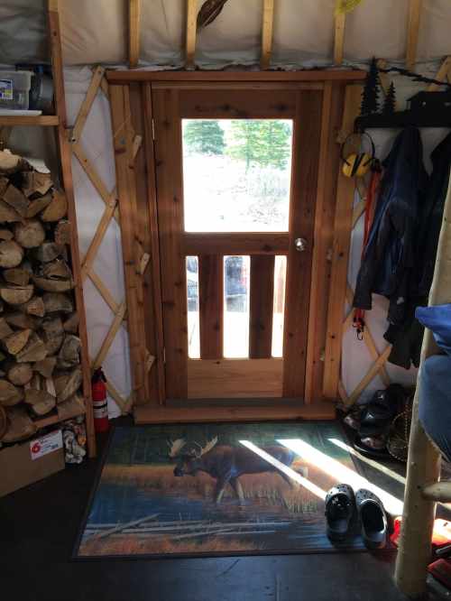 Interior of a yurt with a wooden door, a moose rug, firewood stacked, and shoes by the entrance. Natural light outside.