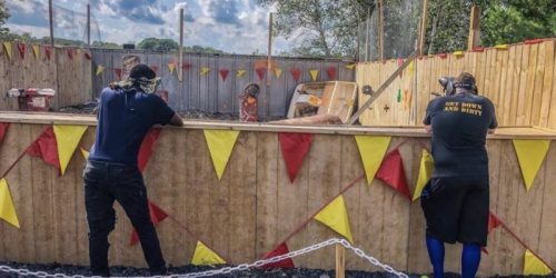 Two people watch from behind a wooden barrier with colorful flags, observing an activity in a fenced area.