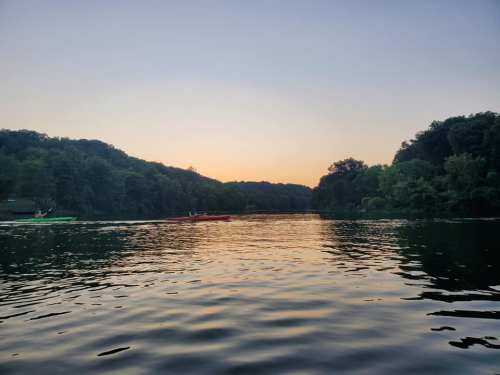 Two kayakers paddle on a calm river at sunset, surrounded by lush greenery and a serene landscape.