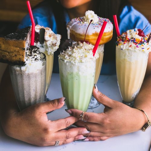A person holds five colorful milkshakes topped with whipped cream, sprinkles, and desserts, showcasing a variety of flavors.