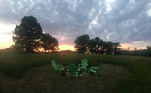 A serene outdoor scene with green chairs arranged around a fire pit, set against a colorful sunset and cloudy sky.