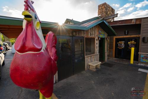A large red rooster statue stands outside a rustic building with a stone chimney and a sign for Flat Creek.