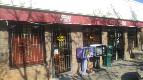 Exterior of a storefront with a maroon awning, featuring multiple windows and a newspaper stand. Signs indicate "Open" and "Hours."