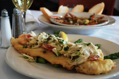 A plate of fried fish topped with shrimp, crab, and coleslaw, served with lemon and asparagus, with bread in the background.