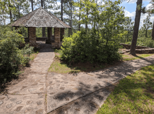 A stone gazebo surrounded by trees and greenery, with a winding path leading through a peaceful outdoor setting.