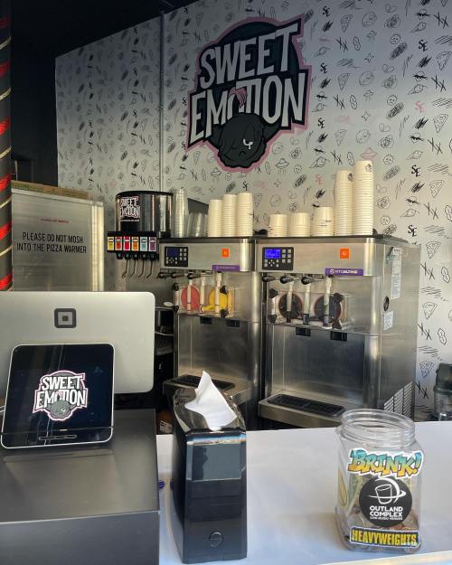 A colorful ice cream shop counter with machines, cups, and a sign reading "Sweet Emotion" on the wall.
