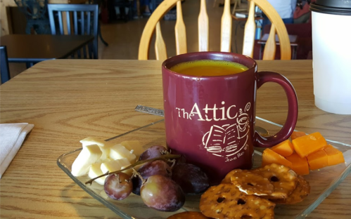 A maroon mug of green tea sits on a table with a plate of grapes, cheese, pretzels, and orange cubes.