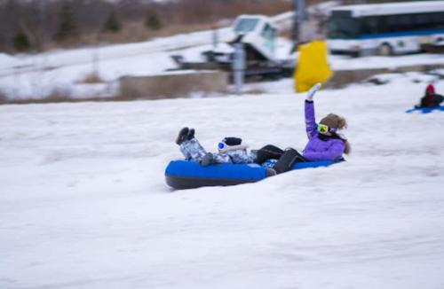 A person in a purple jacket enjoys tubing down a snowy slope, waving with one hand while sitting on a blue inflatable tube.