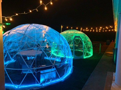 Two illuminated geodesic domes at night, surrounded by string lights, creating a cozy outdoor dining atmosphere.