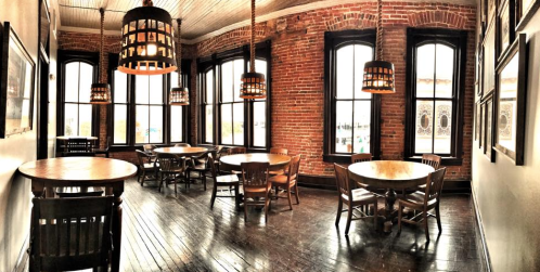 Cozy dining area with wooden tables and chairs, large windows, and exposed brick walls, illuminated by hanging lamps.