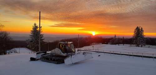 A snow groomer parked on a snowy slope at sunset, with trees and mountains in the background.