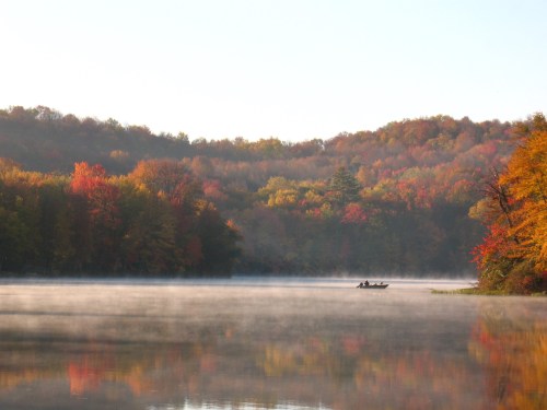 A serene lake surrounded by colorful autumn trees, with a small boat gliding through the misty water.