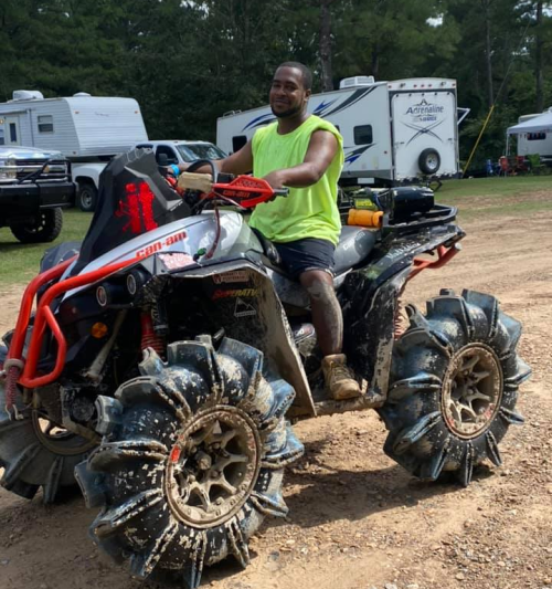 A person in a neon green shirt sits on a muddy ATV in a campsite with trailers in the background.