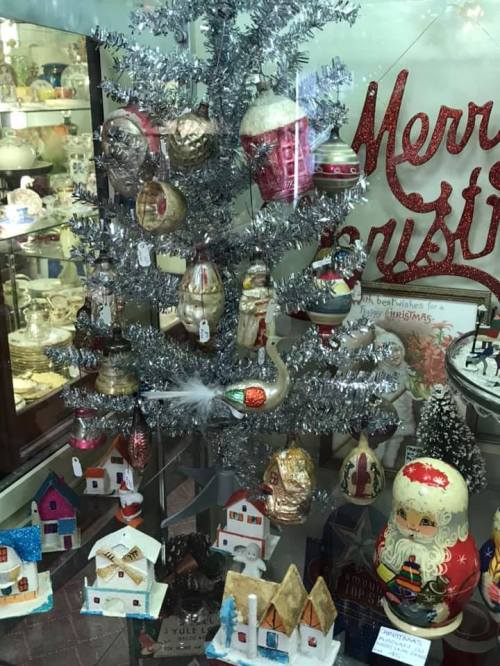 A silver tinsel Christmas tree adorned with vintage ornaments, surrounded by small decorative houses and a Santa figure.