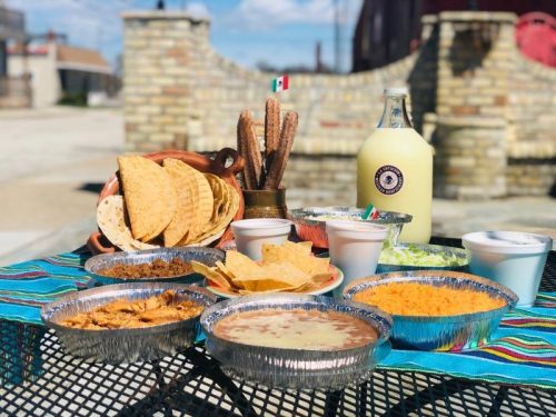 A colorful spread of Mexican food, including tacos, chips, cheese, and drinks, set on a table outdoors.