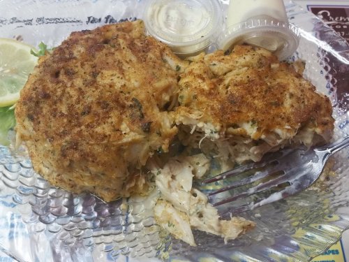 A plate with a large crab cake, partially cut open, accompanied by two small containers of sauce and a lemon wedge.