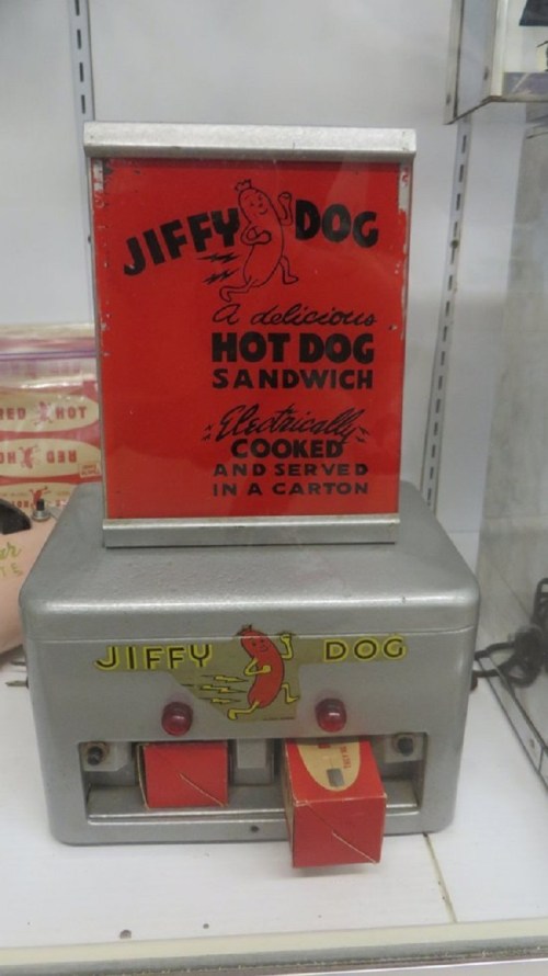 Vintage hot dog vending machine with a red sign reading "Jiffy Dog" and a cartoon hot dog character.