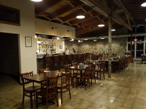 Interior of a rustic restaurant with wooden tables, a bar area, and stone walls, featuring warm lighting and a cozy atmosphere.
