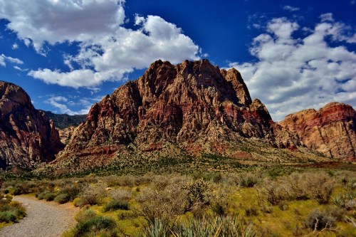 A scenic view of rugged red rock mountains under a blue sky with fluffy clouds, surrounded by desert vegetation.