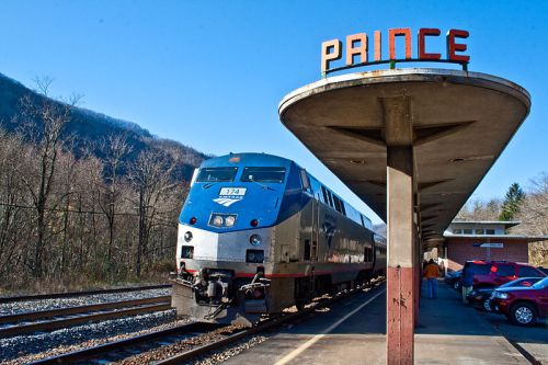 Amtrak train at Prince station, with a vintage sign and passengers waiting on the platform against a mountain backdrop.
