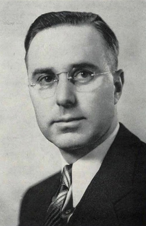 Black and white portrait of a man in a suit and tie, wearing glasses, with a serious expression.