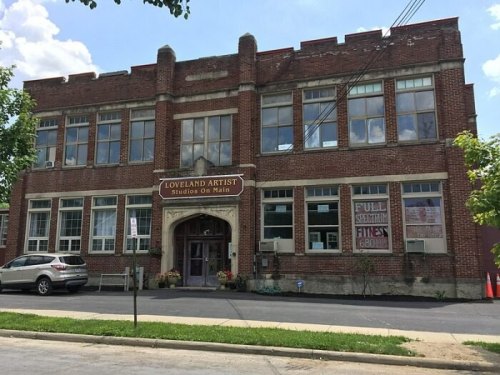 Historic brick building with large windows, labeled "Loveland Artist Studios On Main," surrounded by trees and a parking area.