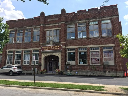 Historic brick building with large windows, labeled "Loveland Artist Studios on Main," surrounded by greenery.