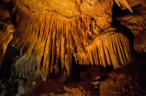Stalactites and stalagmites illuminated in a cave, showcasing intricate rock formations and warm lighting.