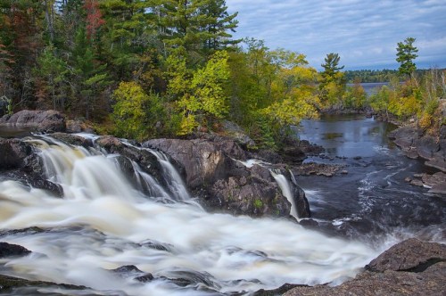 A serene waterfall cascades over rocks, surrounded by vibrant autumn foliage and a calm river under a cloudy sky.