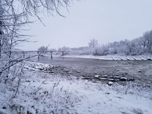 A snowy riverbank scene with a bridge in the background, surrounded by frosted trees and rocks along the water's edge.