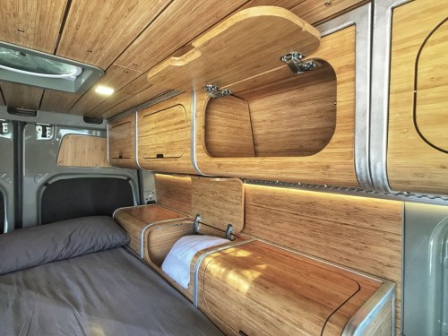 Interior of a van featuring wooden shelves, a bed, and modern lighting, creating a cozy, minimalist space.