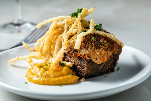 A plated dish featuring crispy fried onions atop a savory meat portion, accompanied by a smooth mustard sauce.