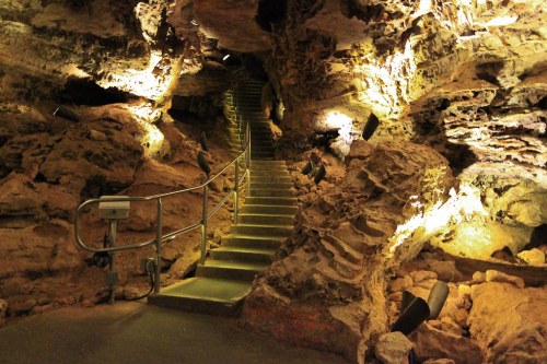 A dimly lit cave interior with a staircase leading upward, surrounded by rocky walls and soft lighting.