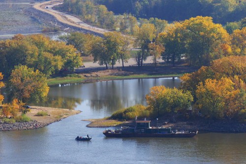 A scenic view of a river with a boat, surrounded by trees in autumn colors and a winding road in the background.