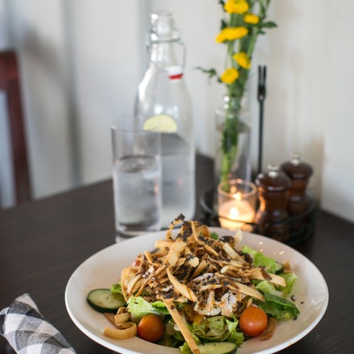 A fresh salad with lettuce, tomatoes, cucumbers, and crispy toppings, alongside a glass of water and a vase of flowers.