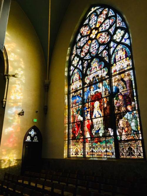 Stained glass window in a church, featuring colorful biblical scenes and light reflections on the wall.