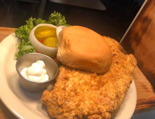 A plate featuring a crispy fried chicken breast, a bun, lettuce, pickles, onions, and a small cup of sour cream.