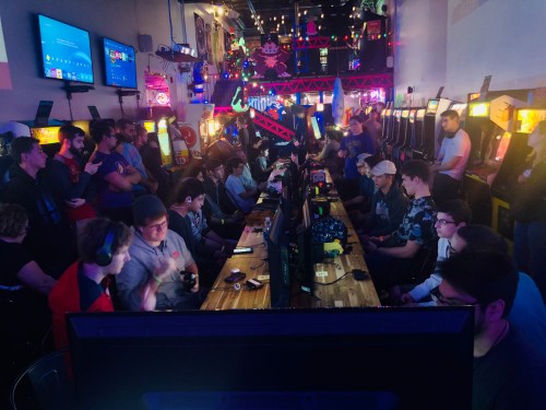 A crowded gaming venue with people playing video games at multiple stations, surrounded by arcade machines and colorful lights.