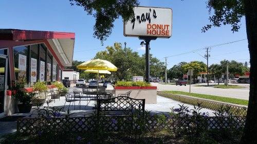 Exterior of Gray's Donut House with outdoor seating, yellow umbrella, and a sign, surrounded by greenery and flowers.