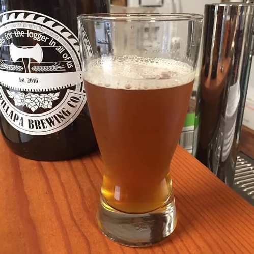 A glass of amber beer with a frothy head, placed on a wooden surface next to a brewing company bottle.