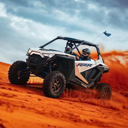 A person drives a Polaris RZR off-road vehicle on a sandy dune, kicking up dust with a blue flag waving.