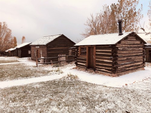 Snow-covered log cabins in a rural setting, surrounded by trees and a grassy area.