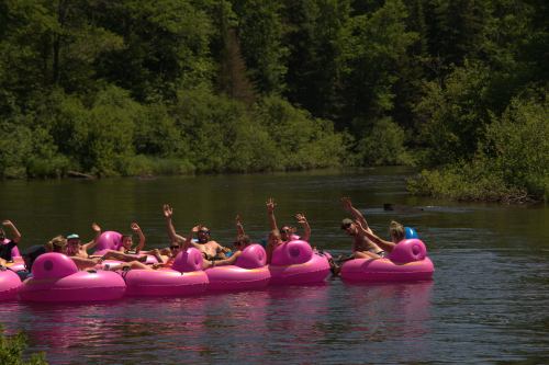A group of people on pink inflatable rafts, waving and enjoying a sunny day on a river surrounded by greenery.