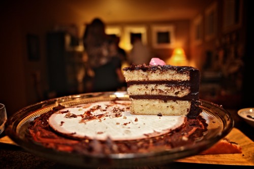 A slice of chocolate cake on a glass plate, with the rest of the cake partially eaten in the background.