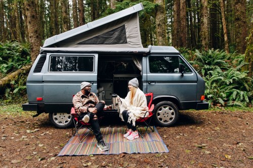 A couple sits in camping chairs by a van in a forest, wrapped in blankets, enjoying a cozy moment together.