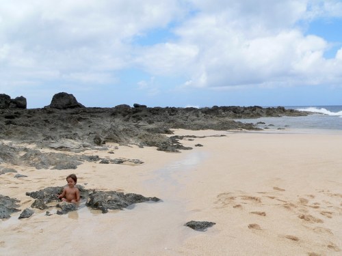 A child sits on a sandy beach near rocky formations, with gentle waves and cloudy skies in the background.