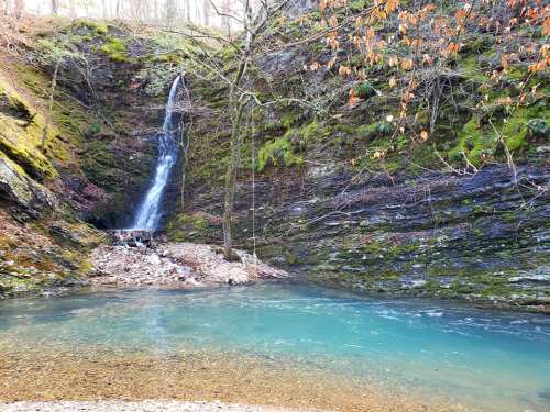 A serene waterfall cascades into a clear blue pool, surrounded by mossy rocks and trees in a tranquil forest setting.