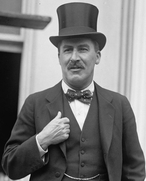 A man in a top hat and bow tie, dressed in formal attire, stands confidently with one hand on his chest.