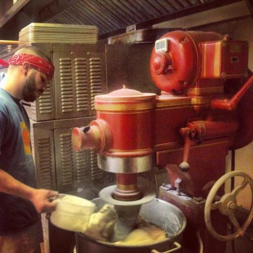 A person in a bandana operates a large red mixer in a kitchen, mixing ingredients in a bowl.
