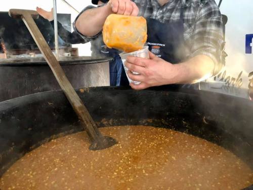 A person ladles a thick, steaming soup from a large pot into a cup at an outdoor event.
